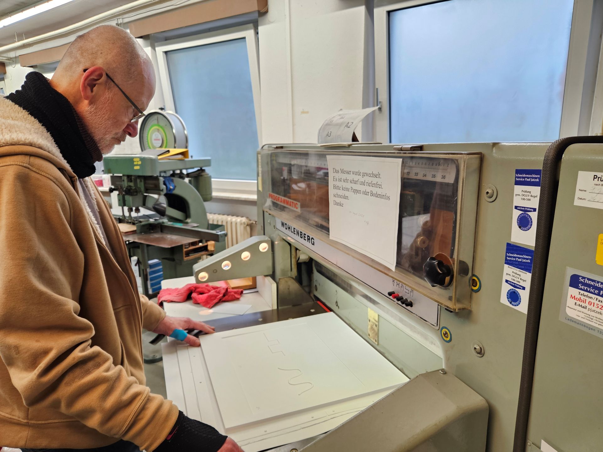 Ein älterer Mann in einer gelben Pulloverjacke steht an einer alten Papierschneidemaschine. Im Hintergrund sind alte Maschinen zu sehen.