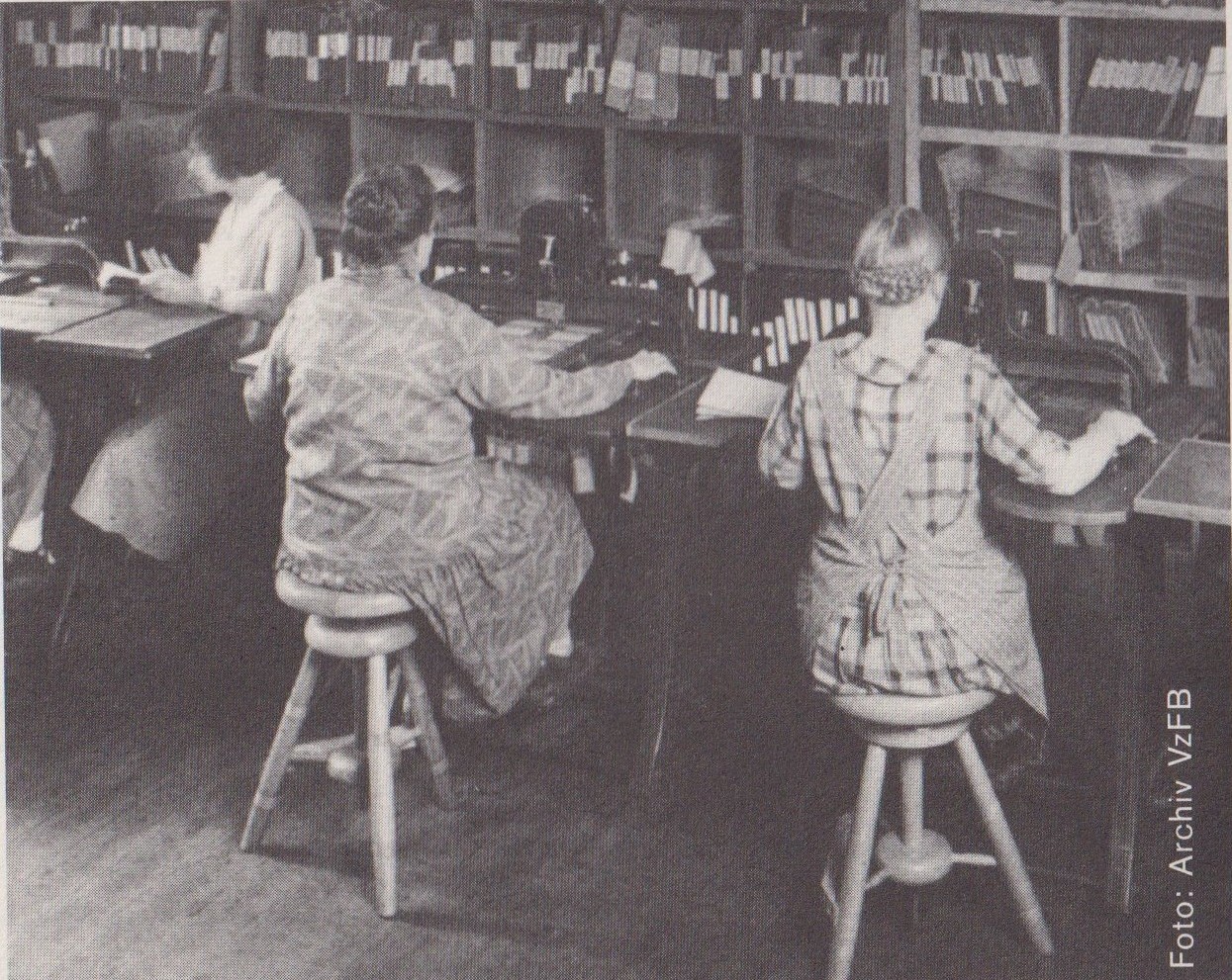 Schwarz-Weiß-Bild. An Arbeitstischen sitzen zwei Frauen, Blick auf die Hinteransicht. Sie übertragen Text in Braille.